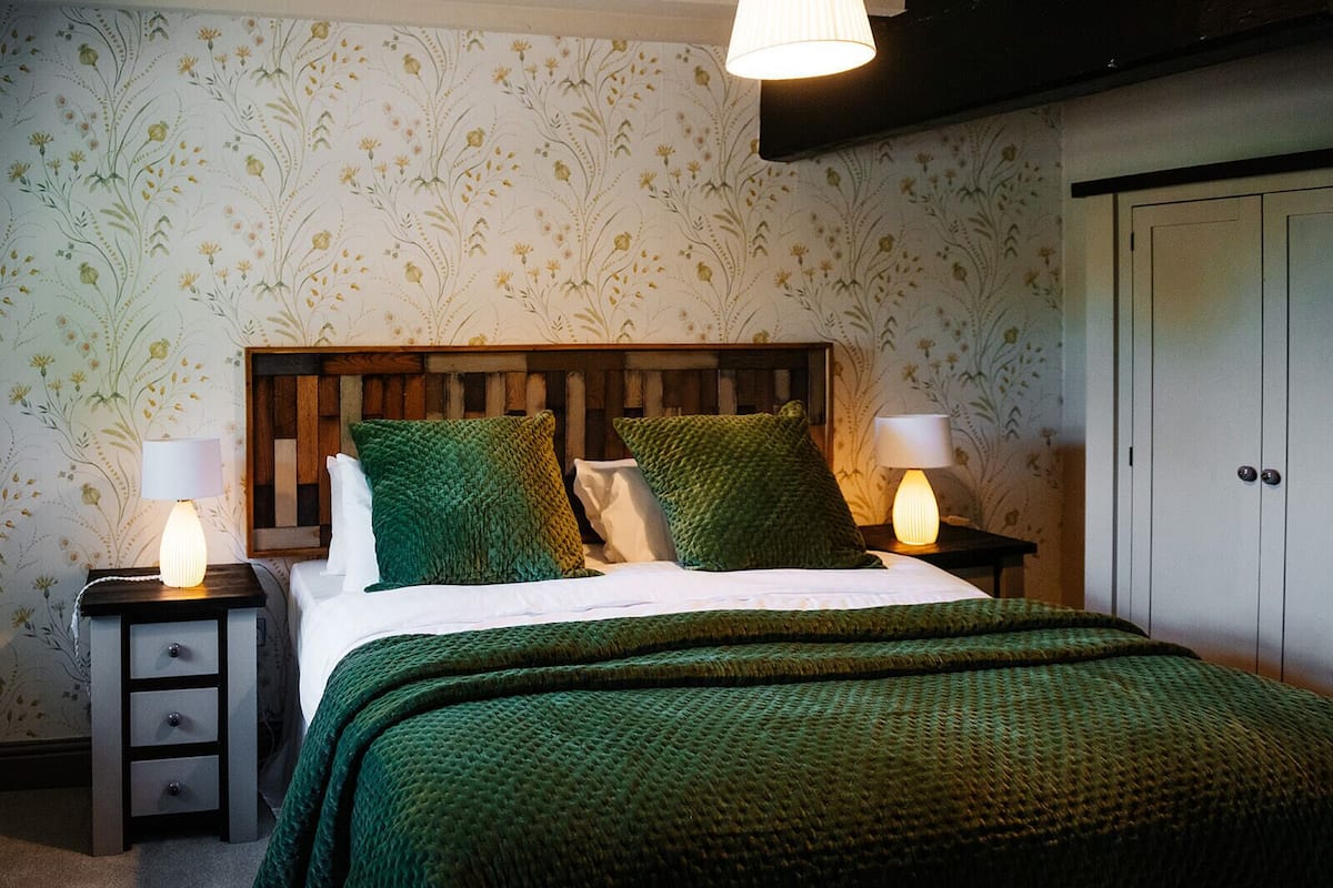 Un dormitorio con cabecera de madera, ropa de cama verde, dos mesitas de noche con lámparas y un armario.