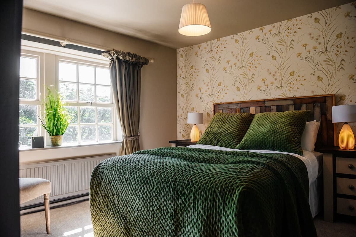 Un dormitorio con cama, dos lámparas de noche, una ventana con cortinas, un radiador y una planta en maceta.