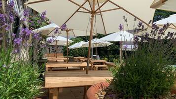 Un patio al aire libre con mesas de madera y grandes sombrillas blancas, rodeado de plantas y flores en macetas.