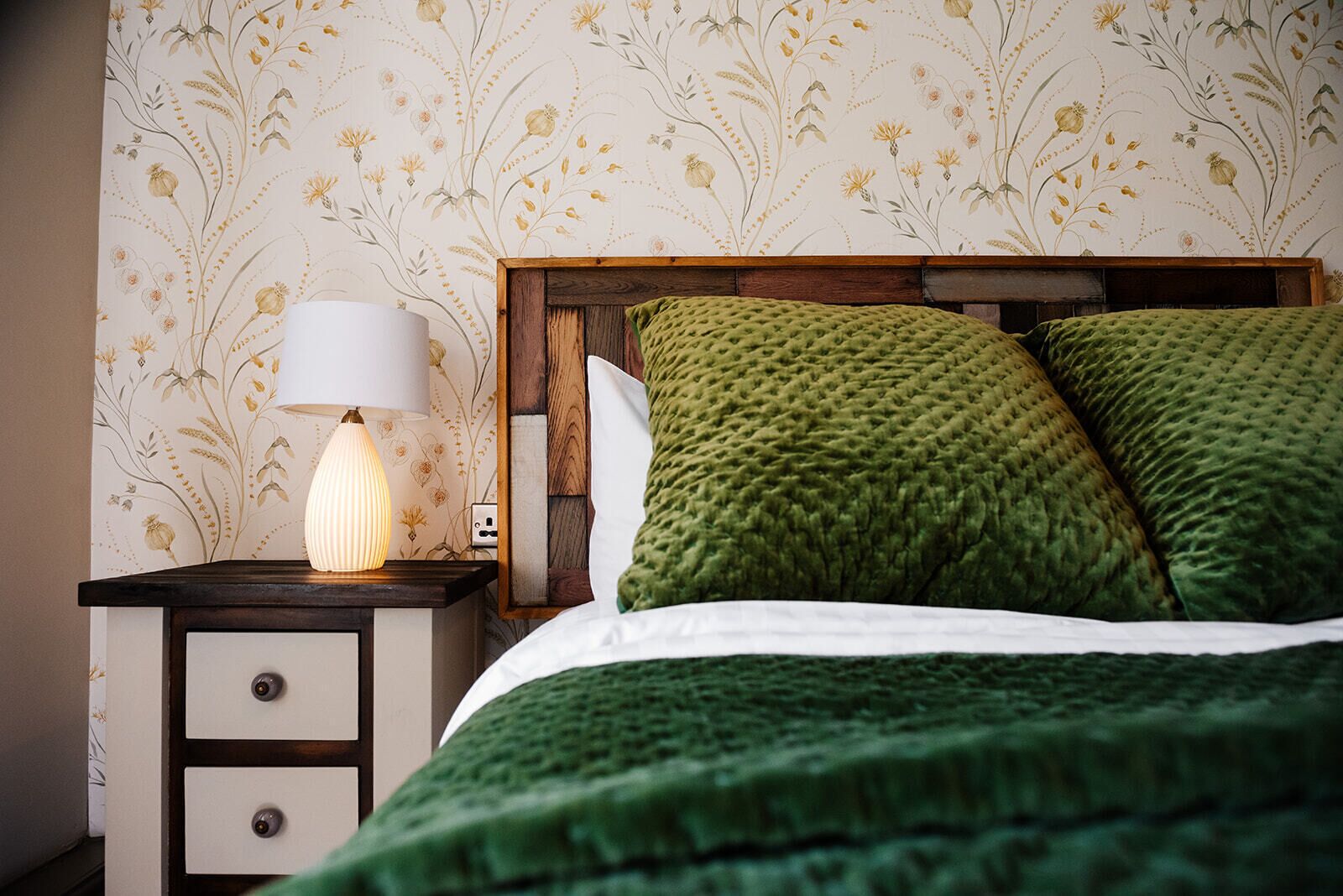A neatly made bed with green bedding, a wooden headboard, and a bedside table with a lamp.