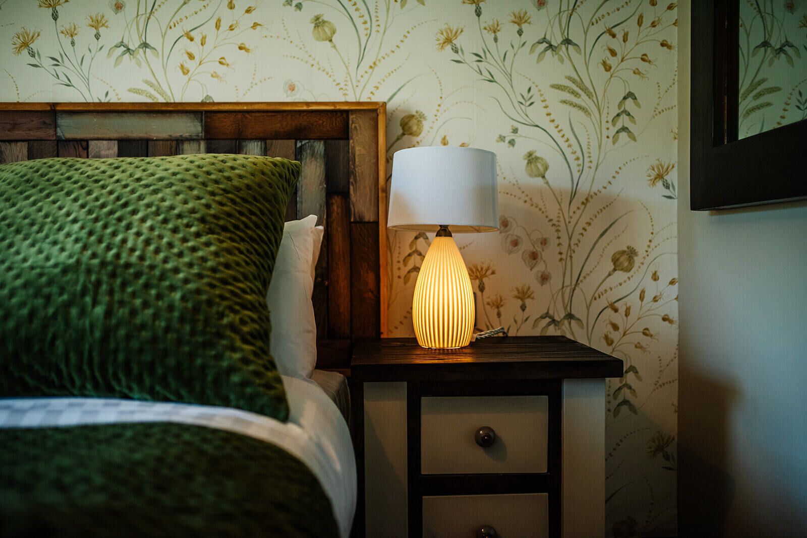 A neatly made bed with a green blanket, a wooden headboard, and a bedside table with a lamp.