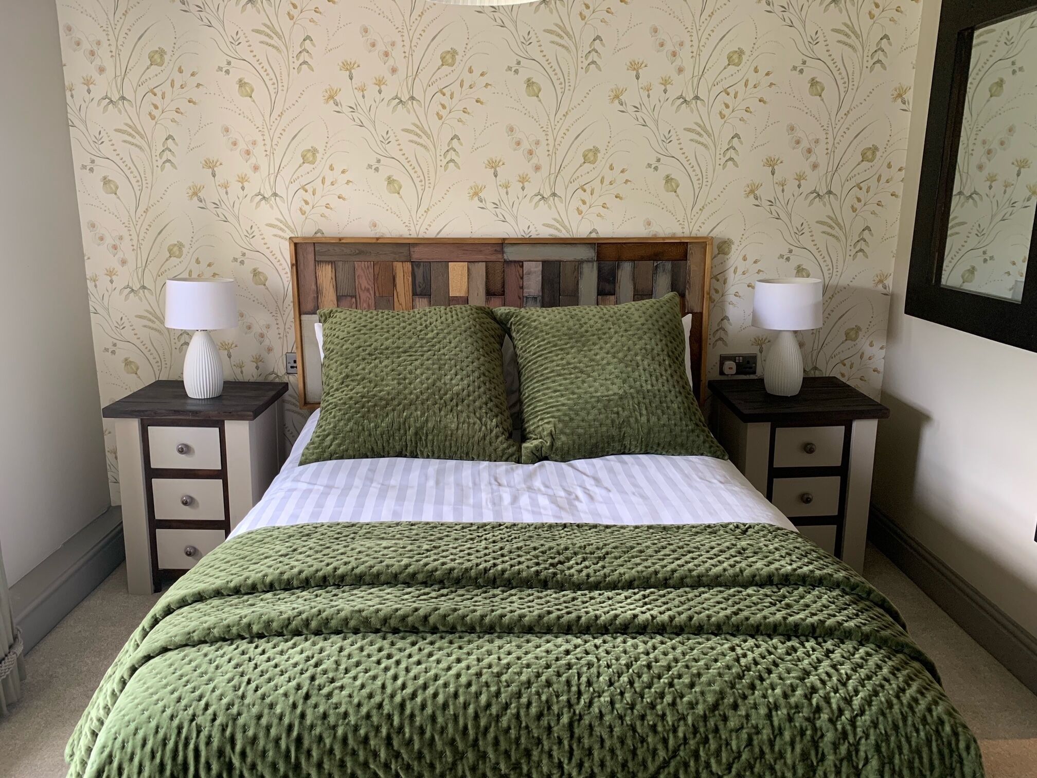 A neatly made bed with a green blanket and pillows, flanked by two bedside tables with lamps, against a floral wallpaper background.
