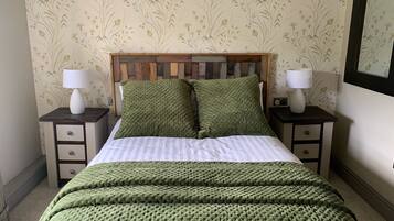 A neatly made bed with a green blanket and pillows, flanked by two bedside tables with lamps, against a floral wallpaper background.
