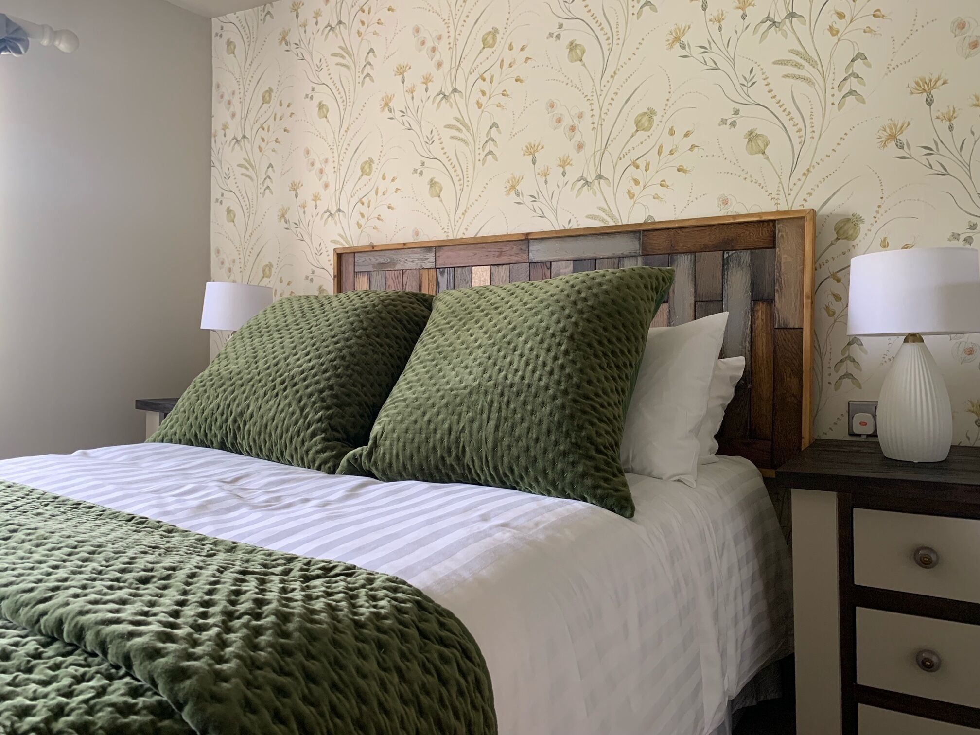 Una cama con manta y almohadas verdes, cabecera de madera, ropa de cama blanca y dos lámparas de noche sobre una mesita.