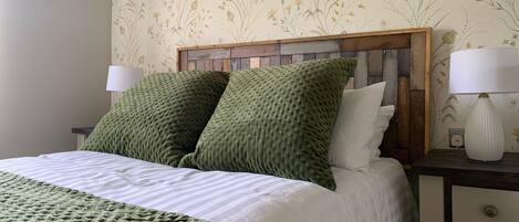Una cama con manta y almohadas verdes, cabecera de madera, ropa de cama blanca y dos lámparas de noche sobre una mesita.