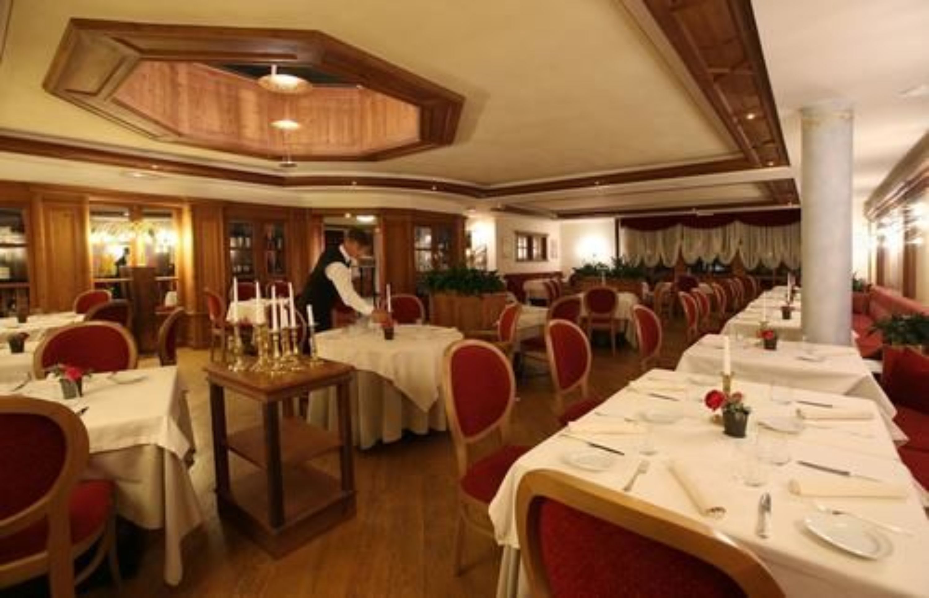 A restaurant with multiple tables set for dining, a waiter attending to guests, and a warm, wooden interior design.