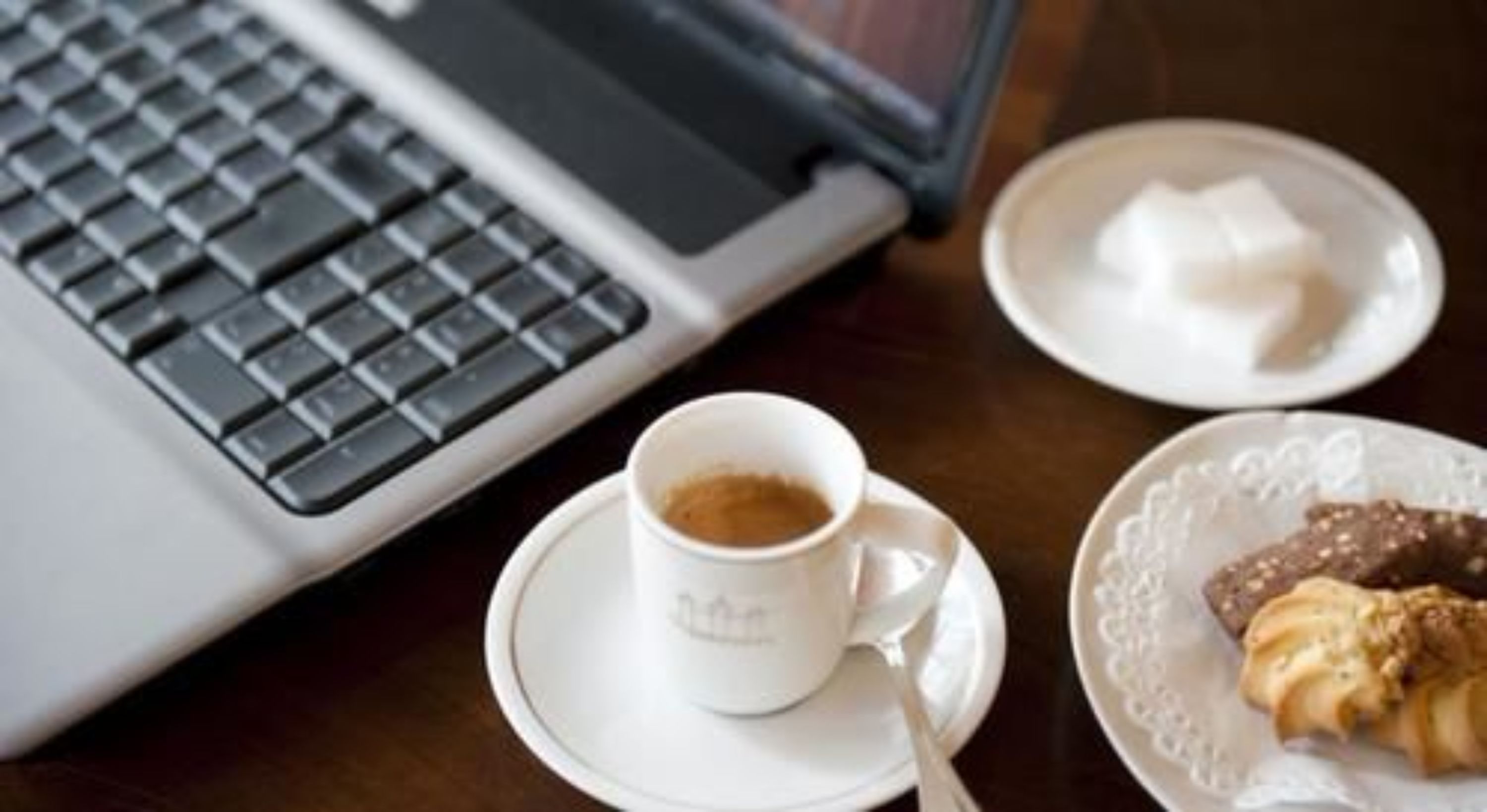 A laptop, a cup of coffee, a plate with a pastry, and a small dish with sugar cubes on a wooden table.