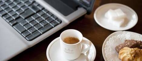 A laptop, a cup of coffee, a plate with a pastry, and a small dish with sugar cubes on a wooden table.