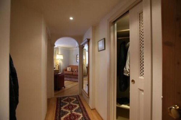 A narrow hallway with a patterned rug, a door leading to a room, and a mirror reflecting a room with a lamp and a sofa.