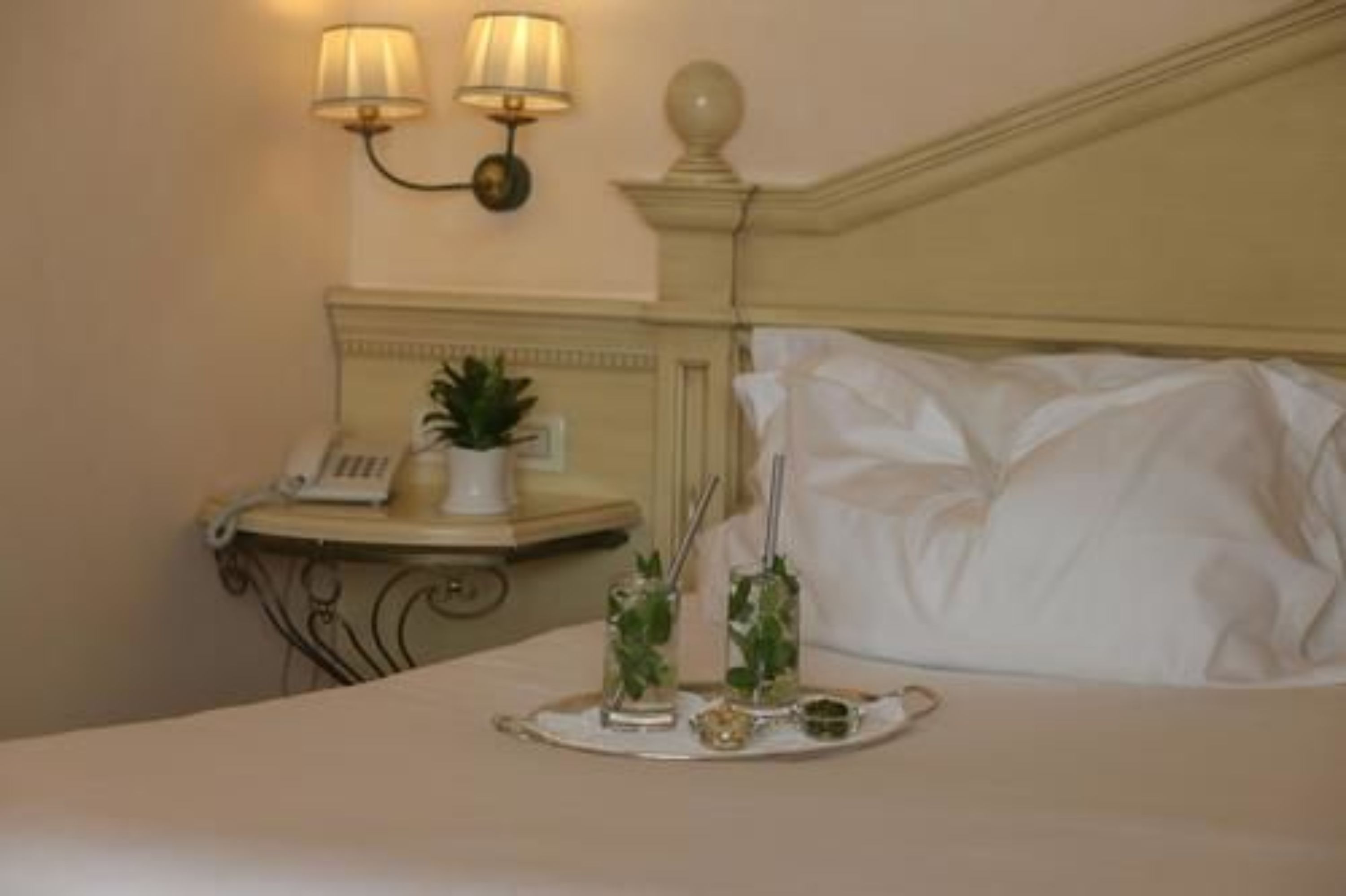 Un letto rifatto con lenzuola bianche, un comodino con una lampada, un telefono e un vassoio con una bevanda e una piccola pianta.