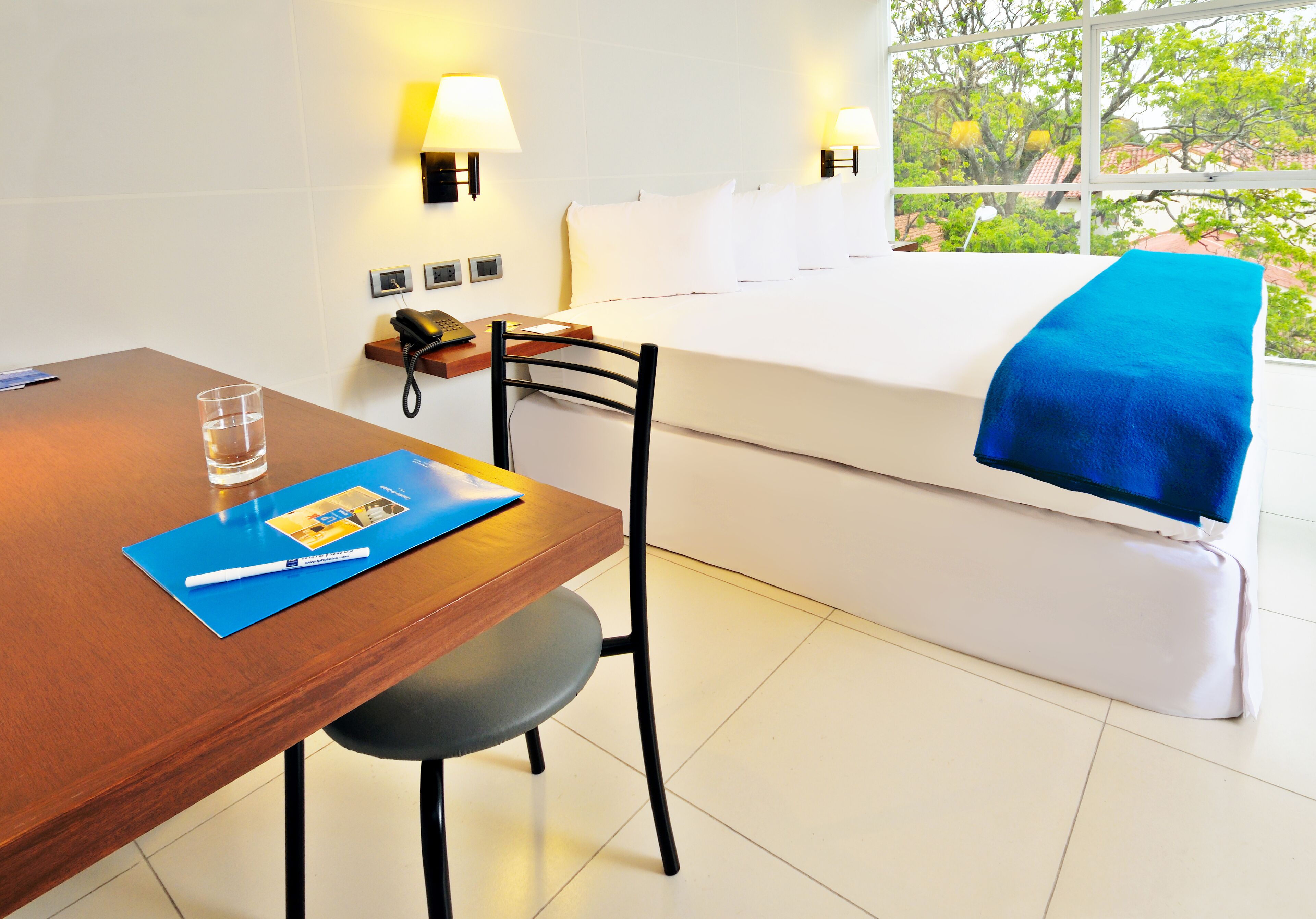 Habitación de hotel con un escritorio de madera, una silla, una cama con manta azul, una mesita de noche con lámpara y una ventana con vistas a la vegetación.