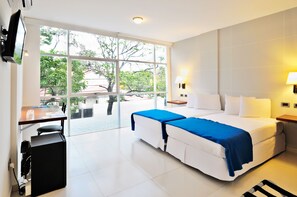 A hotel room with a large window, a desk with a chair, a bed with white bedding and a blue accent, and a TV mounted on the wall.