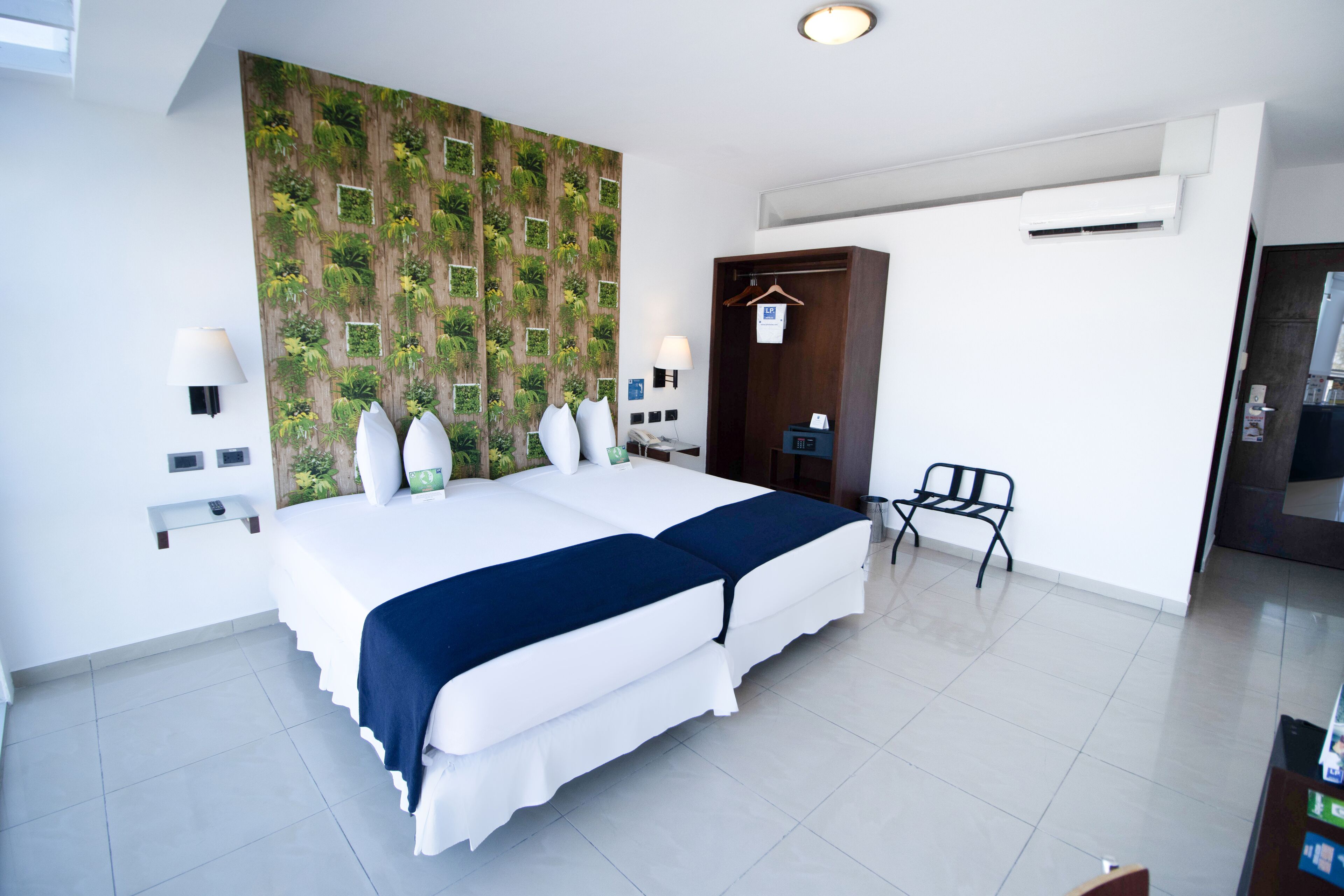 A modern hotel room with a large bed, a wooden wall panel with greenery, a desk with a chair, and a wardrobe.