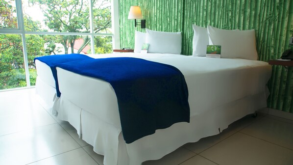 A bed with white bedding and a blue throw, a bedside lamp, and a view of greenery through a window.