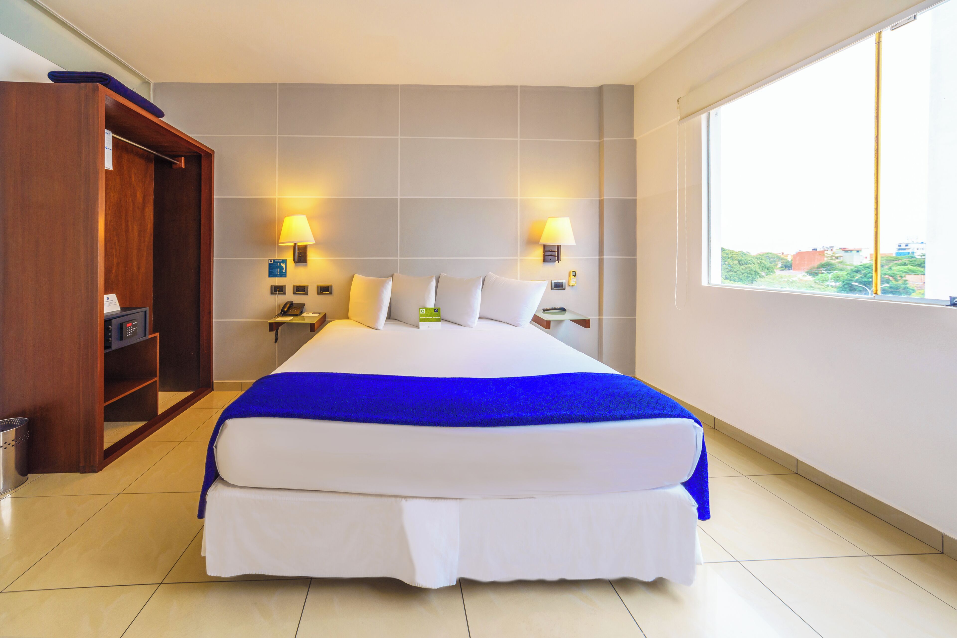 A hotel room with a large bed, two bedside lamps, a wooden wardrobe, and a window with a city view.