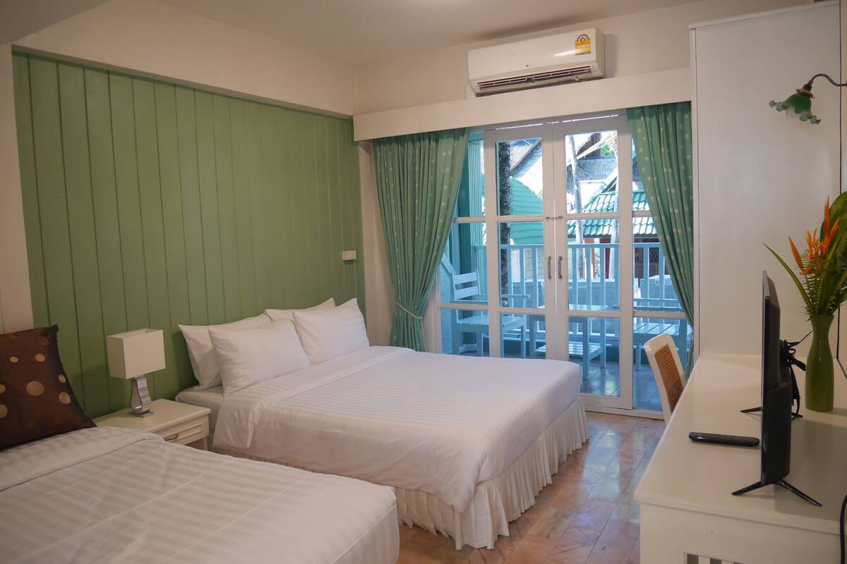 A hotel room with two beds, a desk, a chair, an air conditioner, and a view of a balcony with outdoor furniture.