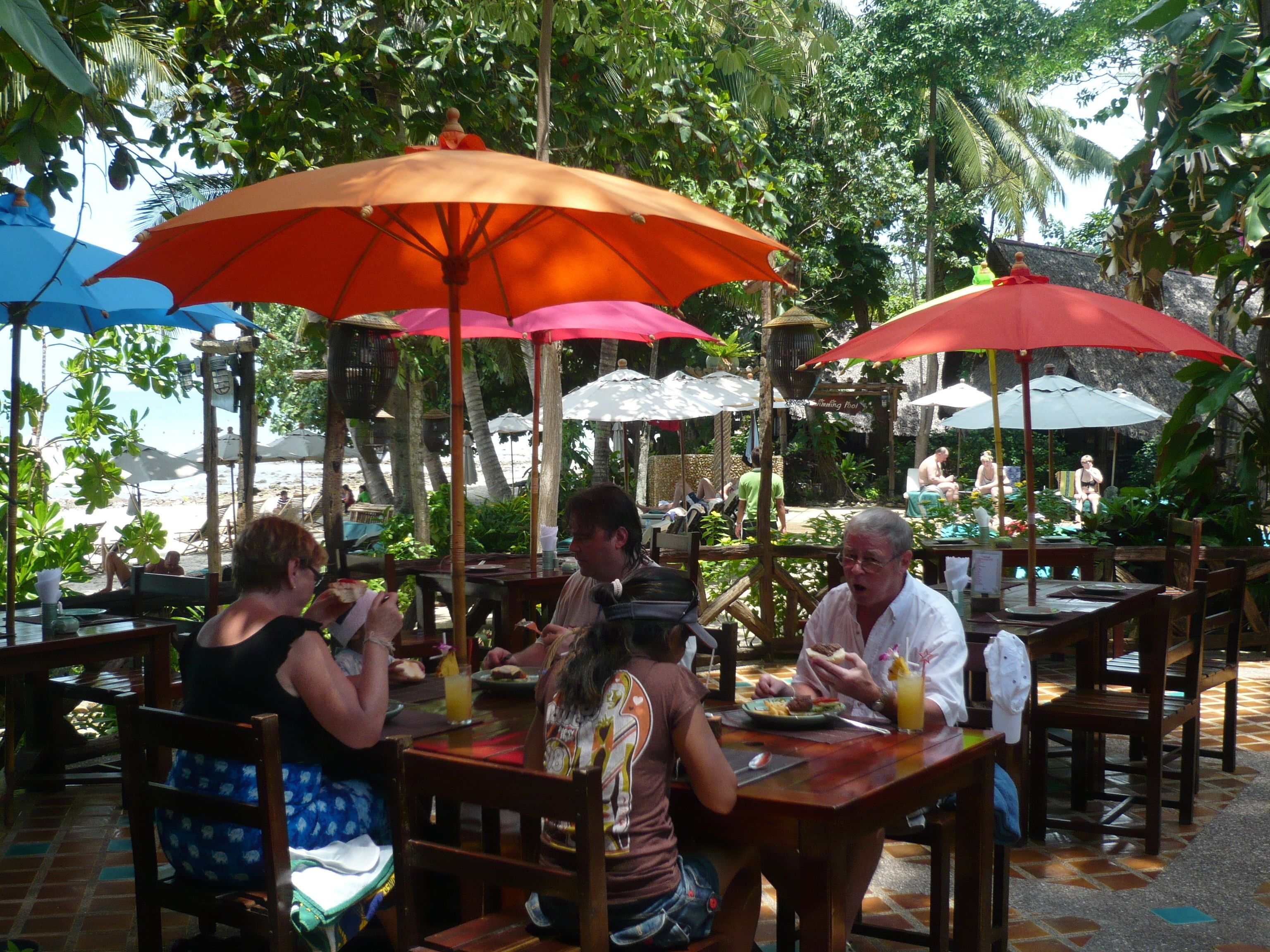 An outdoor dining area with tables and chairs under umbrellas, surrounded by lush greenery and a view of the beach.