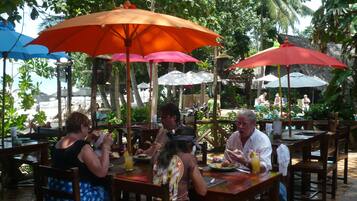 An outdoor dining area with tables and chairs under umbrellas, surrounded by lush greenery and a view of the beach.