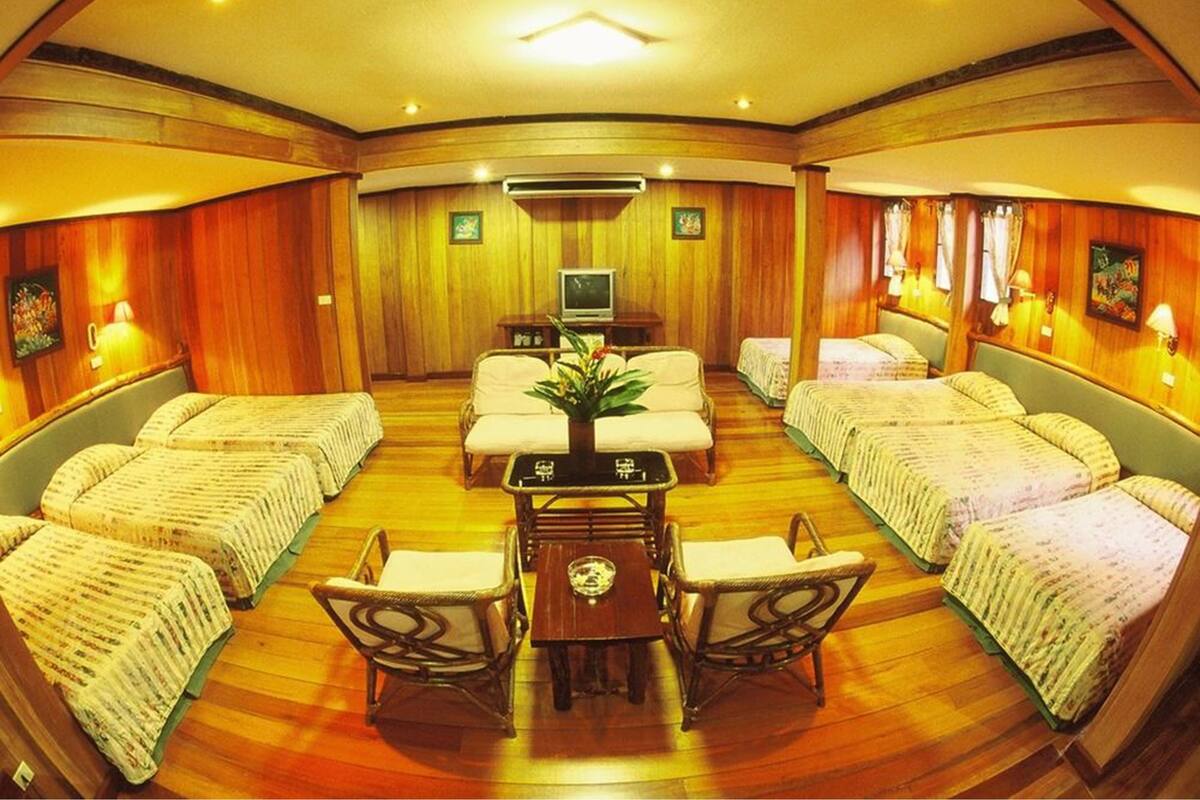 A room with four beds arranged in a semi-circle, a central table with chairs, a television, and wall-mounted lamps.