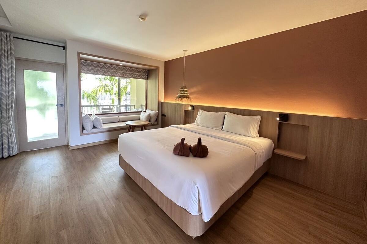 A modern hotel room with a large bed, a seating area by the window, and a view of greenery outside.