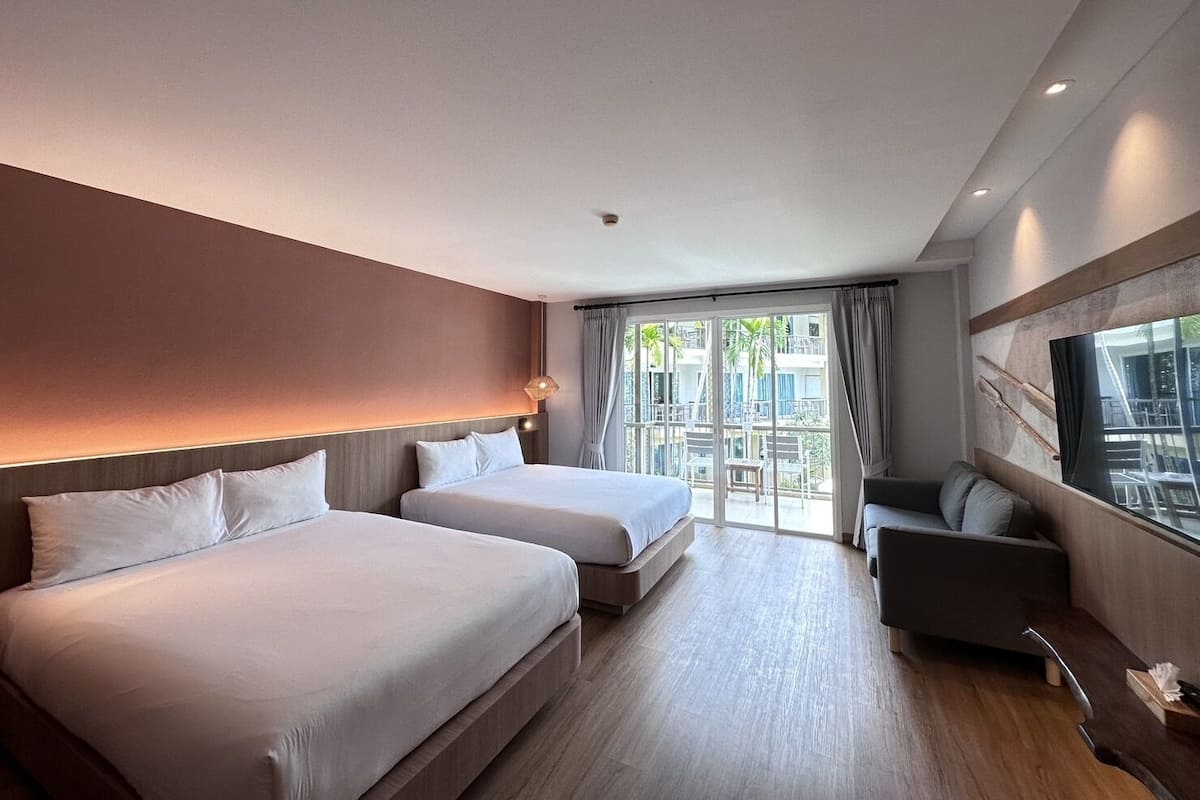 A hotel room with two beds, a sofa, a TV, and a balcony with a view of palm trees.