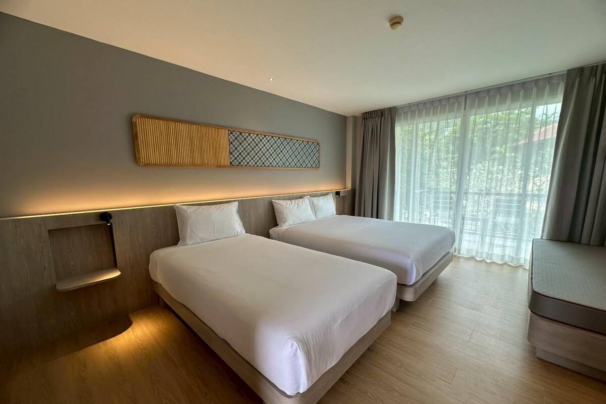A hotel room with two beds, a wooden floor, a large window with curtains, and a wall-mounted light fixture.