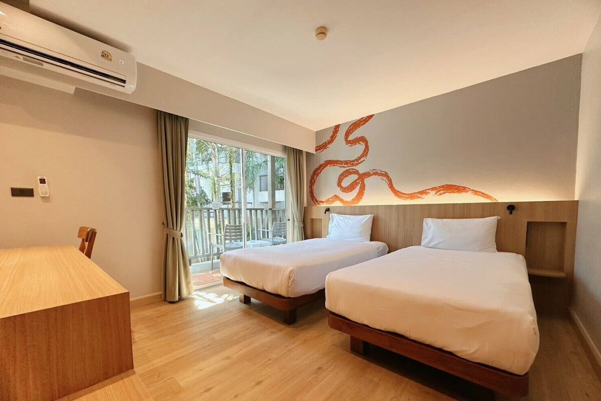 A hotel room with two beds, a wooden desk, a balcony with a view, and an air conditioning unit.