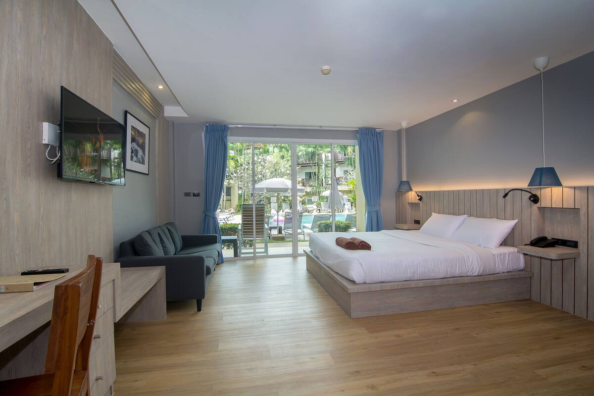 A modern hotel room with a bed, desk, and seating area, featuring a large window and a view of the outdoors.