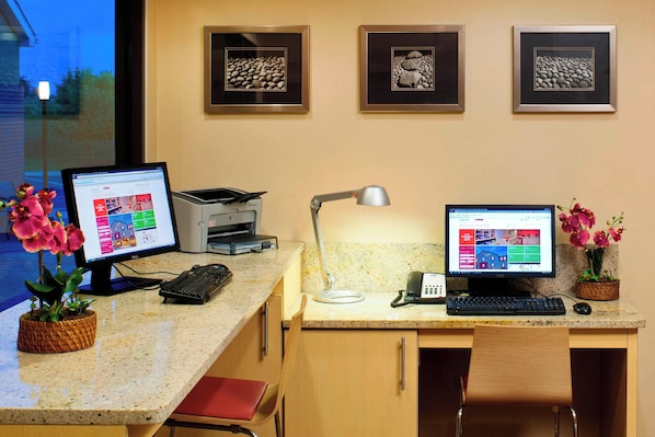 Ein Schreibtisch mit zwei Bildschirmen, einer Tastatur, einer Lampe und einem Telefon. An der Wand hängen gerahmte Bilder und auf dem Schreibtisch steht ein Blumentopf.