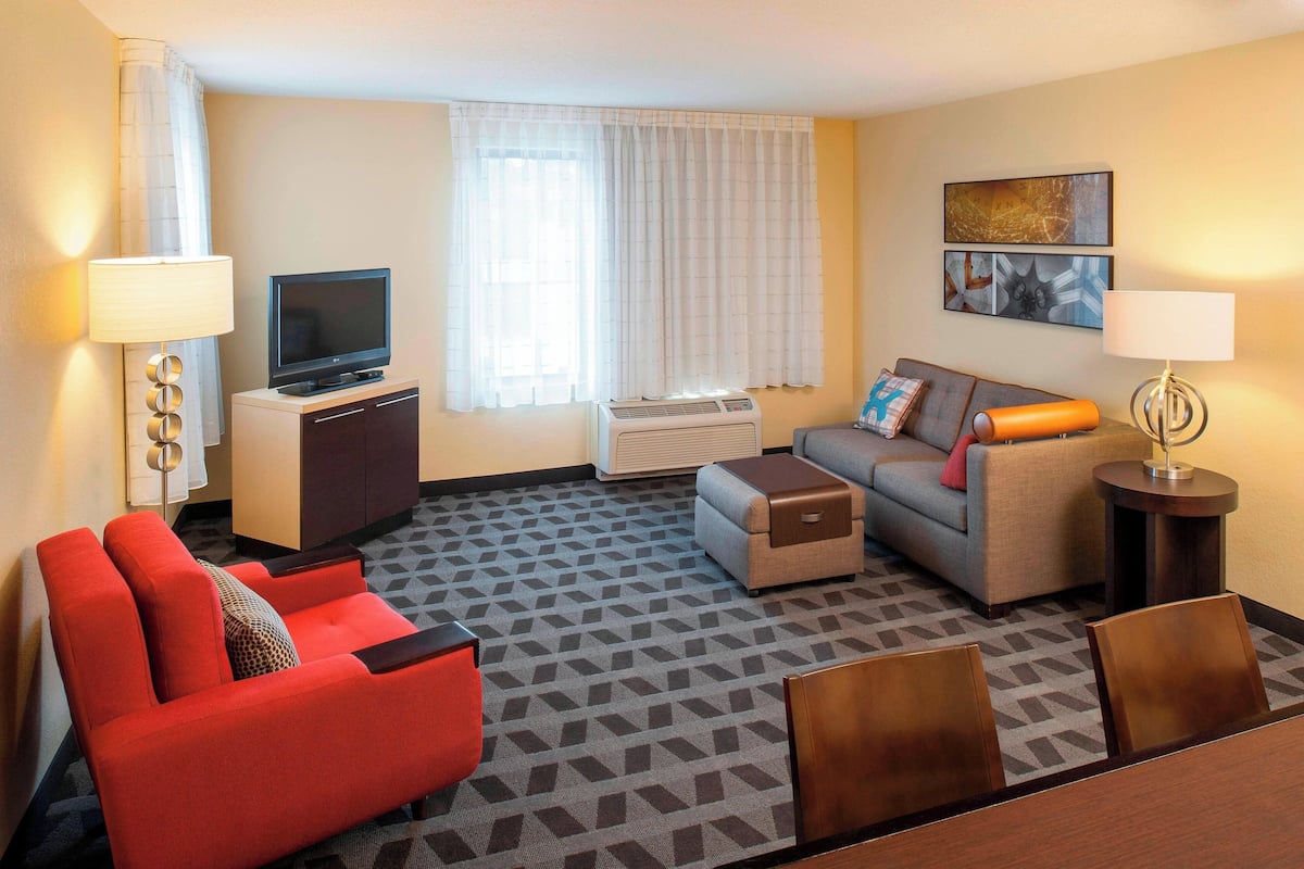 Un cuarto de hotel con un sofá rojo, un sofá gris, un televisor sobre un soporte, una mesita, una lámpara y un cuadro en la pared.