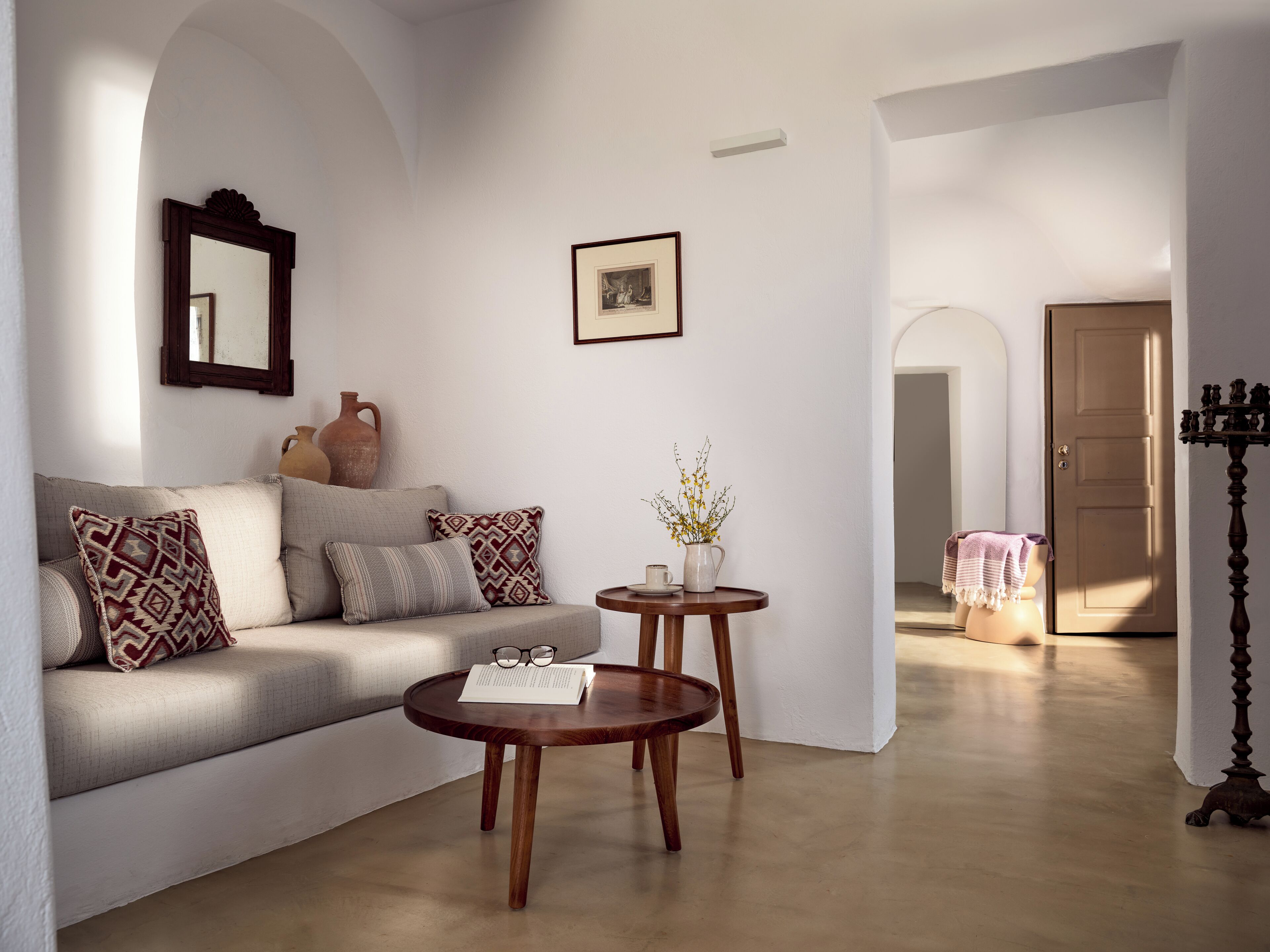 Una sala de estar con un sofá, dos mesitas auxiliares, un espejo, un cuadro enmarcado y una puerta que da a otra habitación.