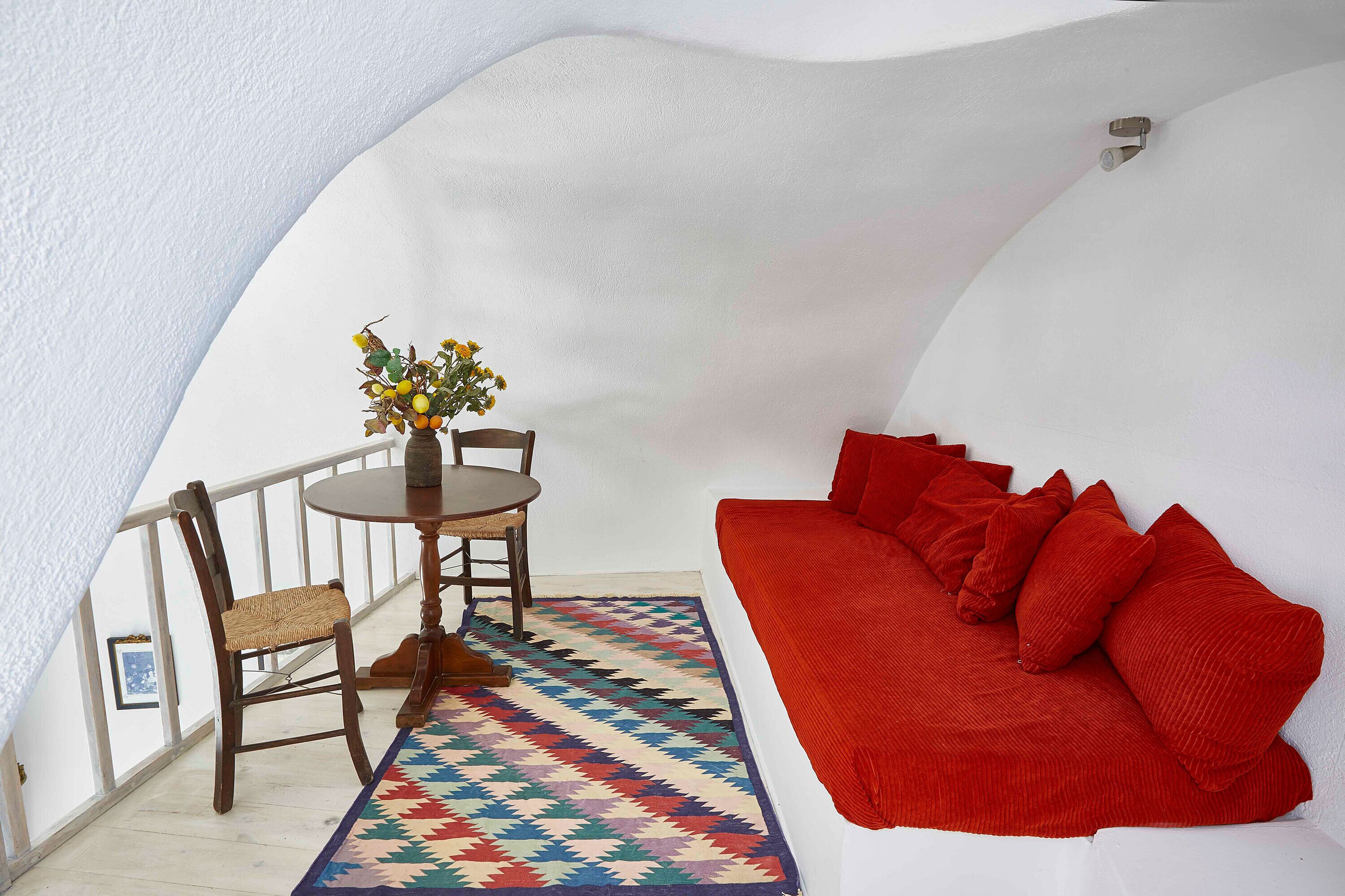A cozy room with a red sofa, a wooden chair, a small table with a vase of flowers, and a colorful rug.