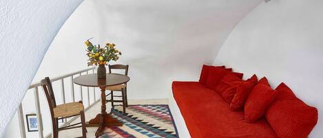 A cozy room with a red sofa, a wooden chair, a small table with a vase of flowers, and a colorful rug.