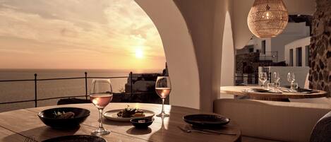 Una mesa de comedor con platos, cubiertos y vasos, con vistas a la puesta de sol sobre el mar.