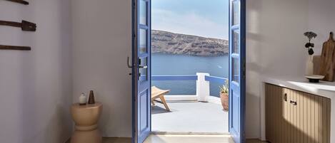 A bedroom with a view of a sea and mountains, a wooden bench, a small table with a vase, and a bed with a patterned cover.