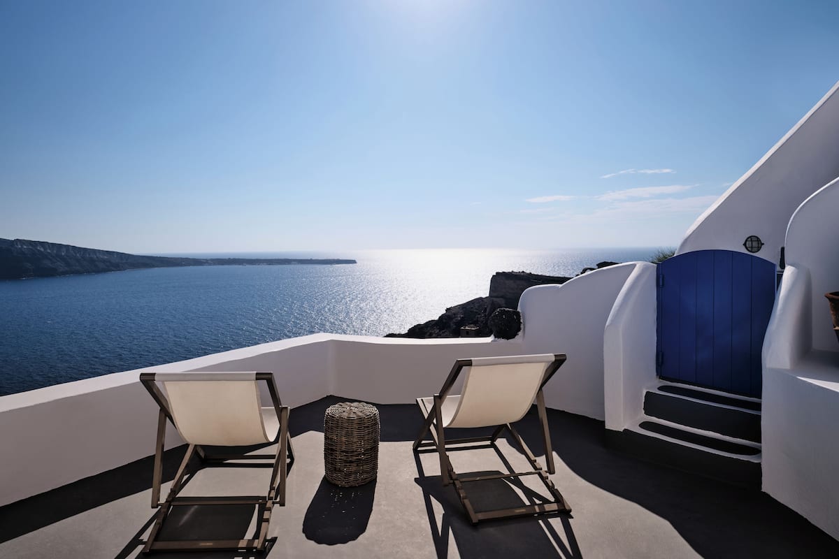 En balkon med to stole og en fletkurv, med udsigt over kysten, en klar himmel og et roligt hav.