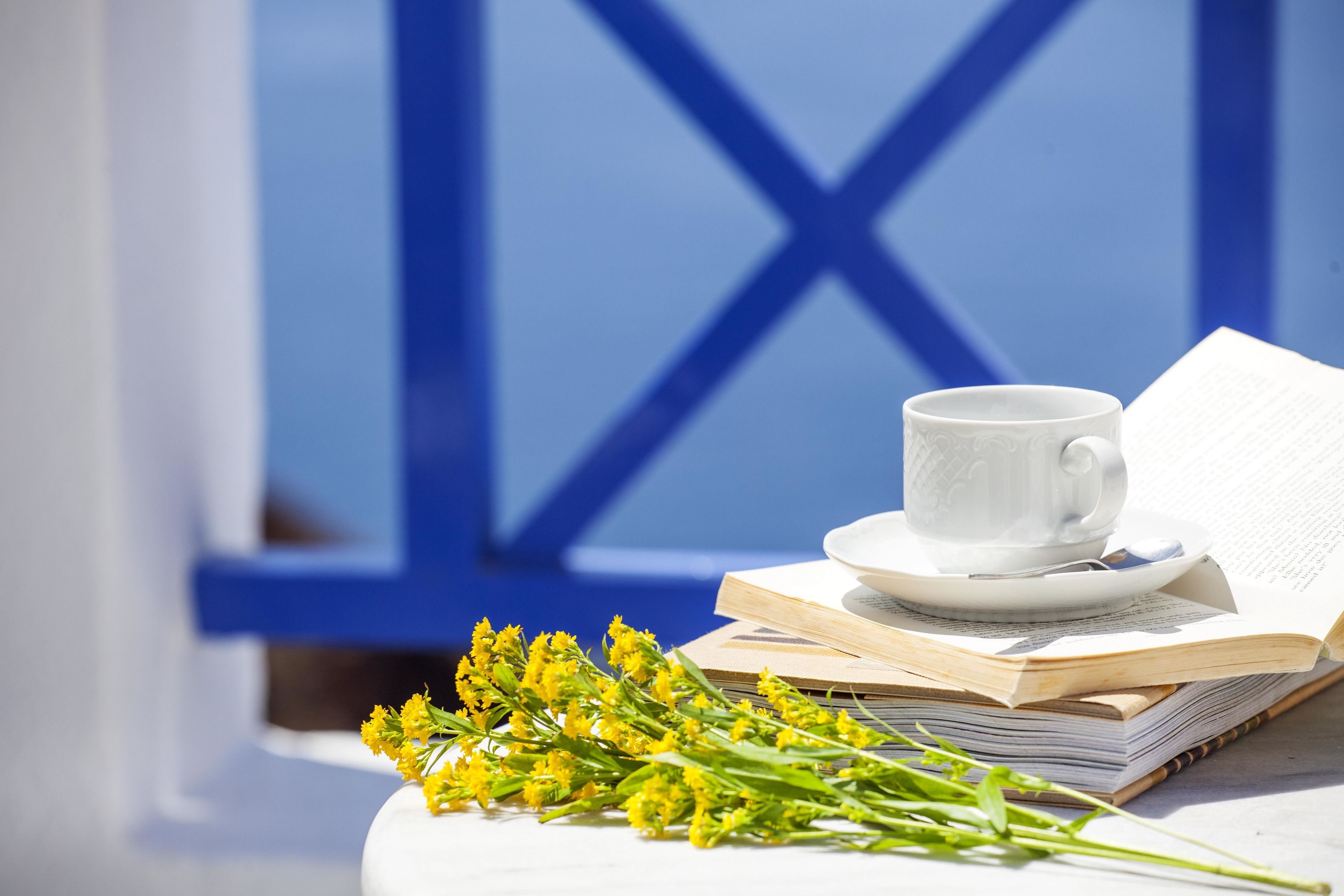 Een witte kop op een schotel met een boek en gele bloemen op een tafel.