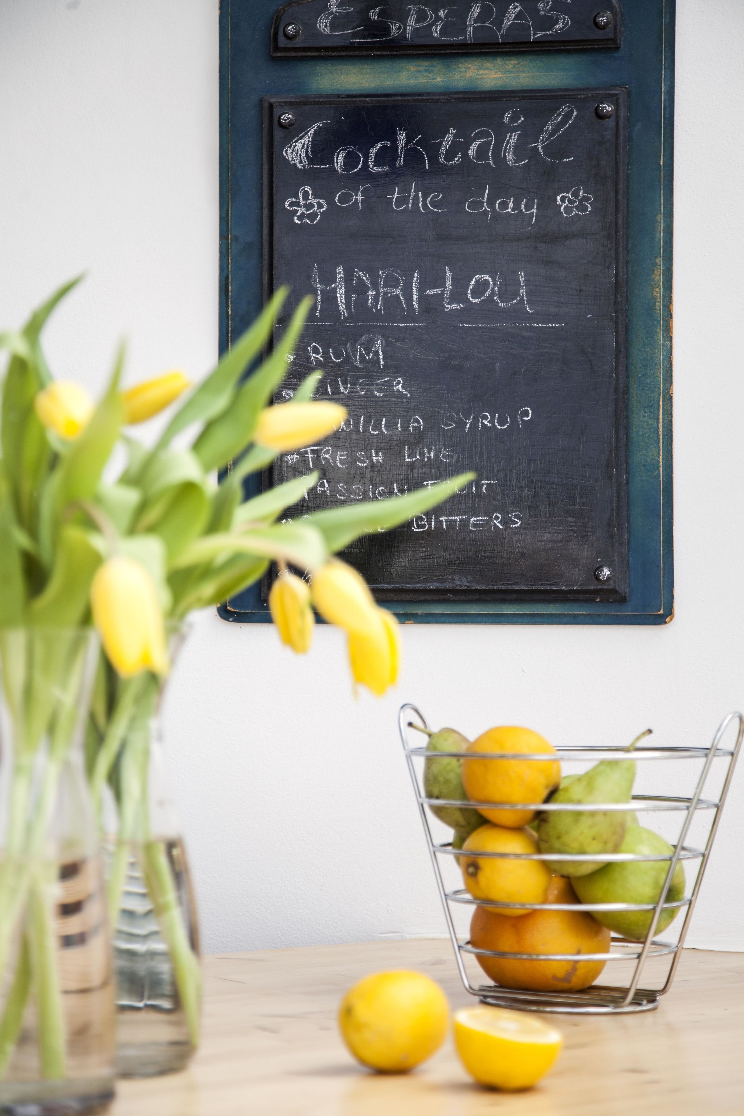 A chalkboard menu listing a cocktail recipe, a vase with yellow flowers, and a fruit basket on a wooden table.