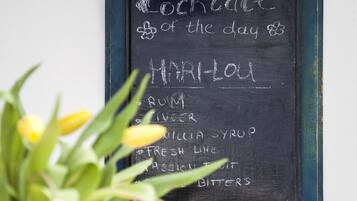 A chalkboard menu listing a cocktail recipe, a vase with yellow flowers, and a fruit basket on a wooden table.