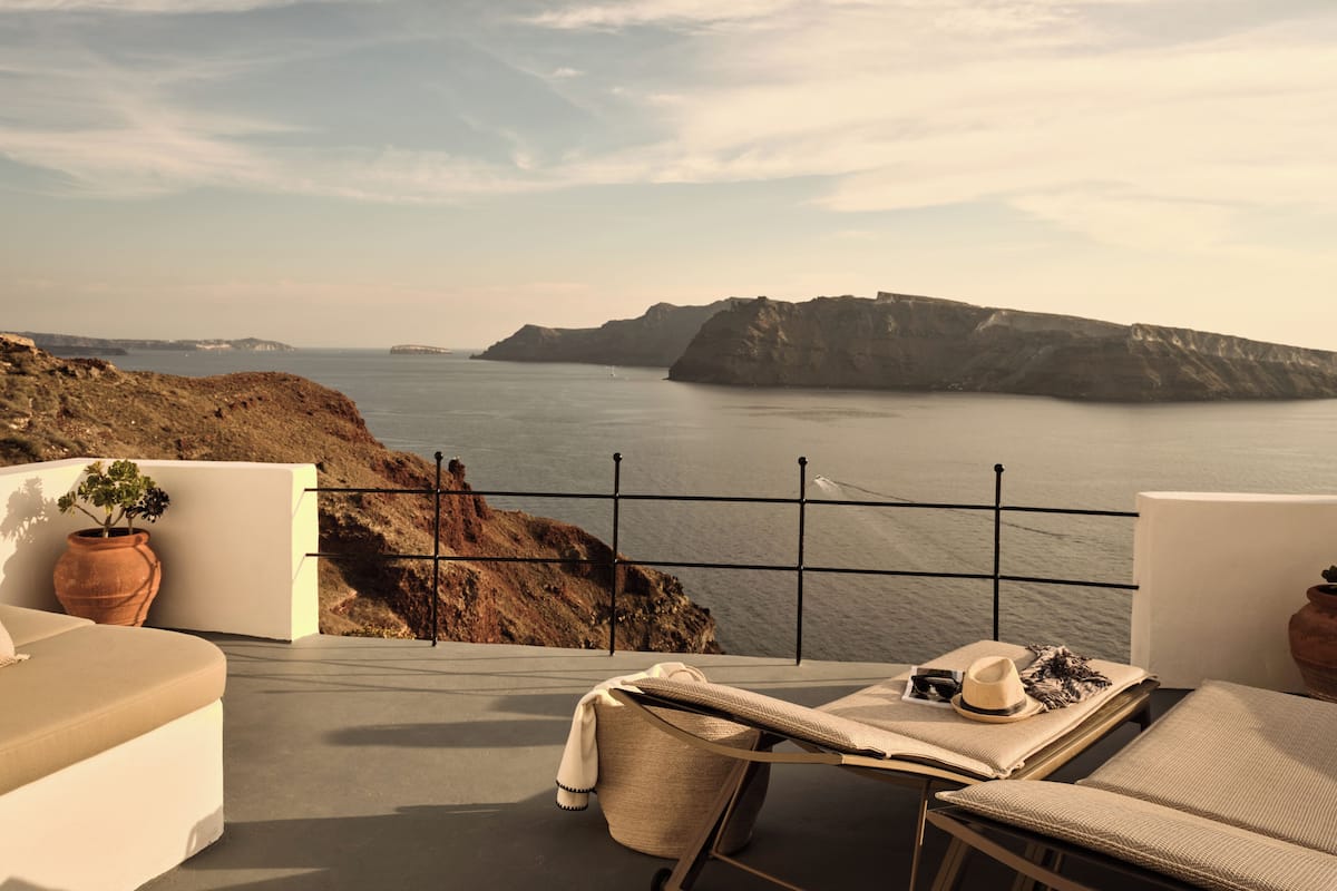 Balkon med udsigt over hav og klipper, en liggestol og et bord med hat og solbriller.