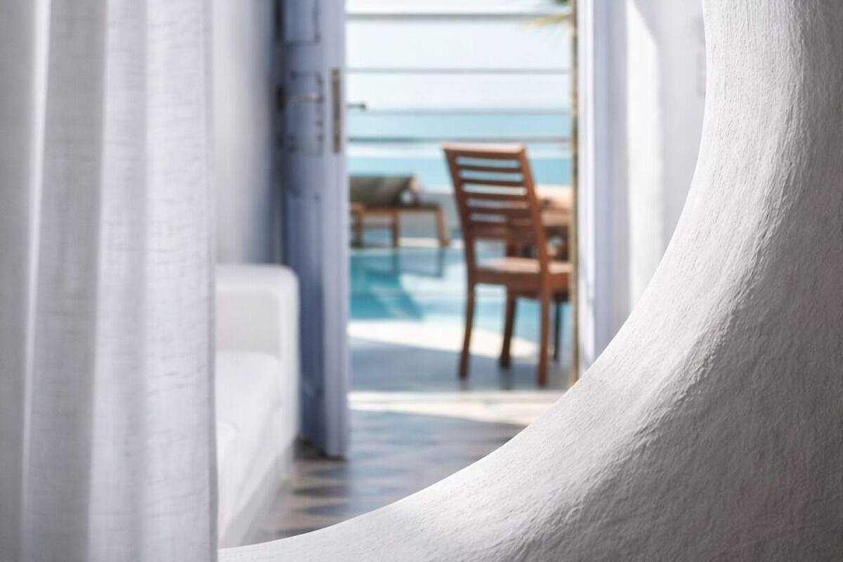 A view through a white, curved architectural feature to a balcony with a pool and chairs, overlooking a clear sky and palm tree.