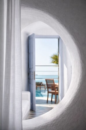Vista a través de un elemento arquitectónico blanco y curvo hacia un balcón con piscina y sillas, con vista a un cielo despejado y una palmera.