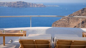 Terraza frente al mar con sillones de madera y una mesa, con vistas a un mar azul cristalino y acantilados rocosos.