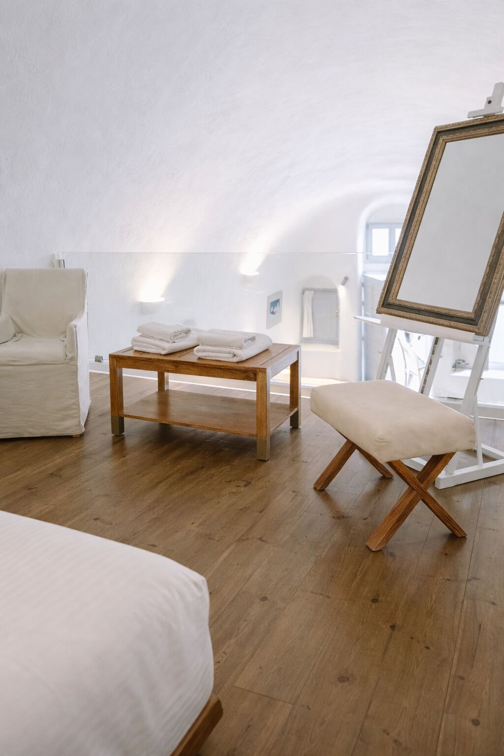 A room with a wooden floor, a white armchair, a wooden table with folded towels, and a large mirror on a stand.