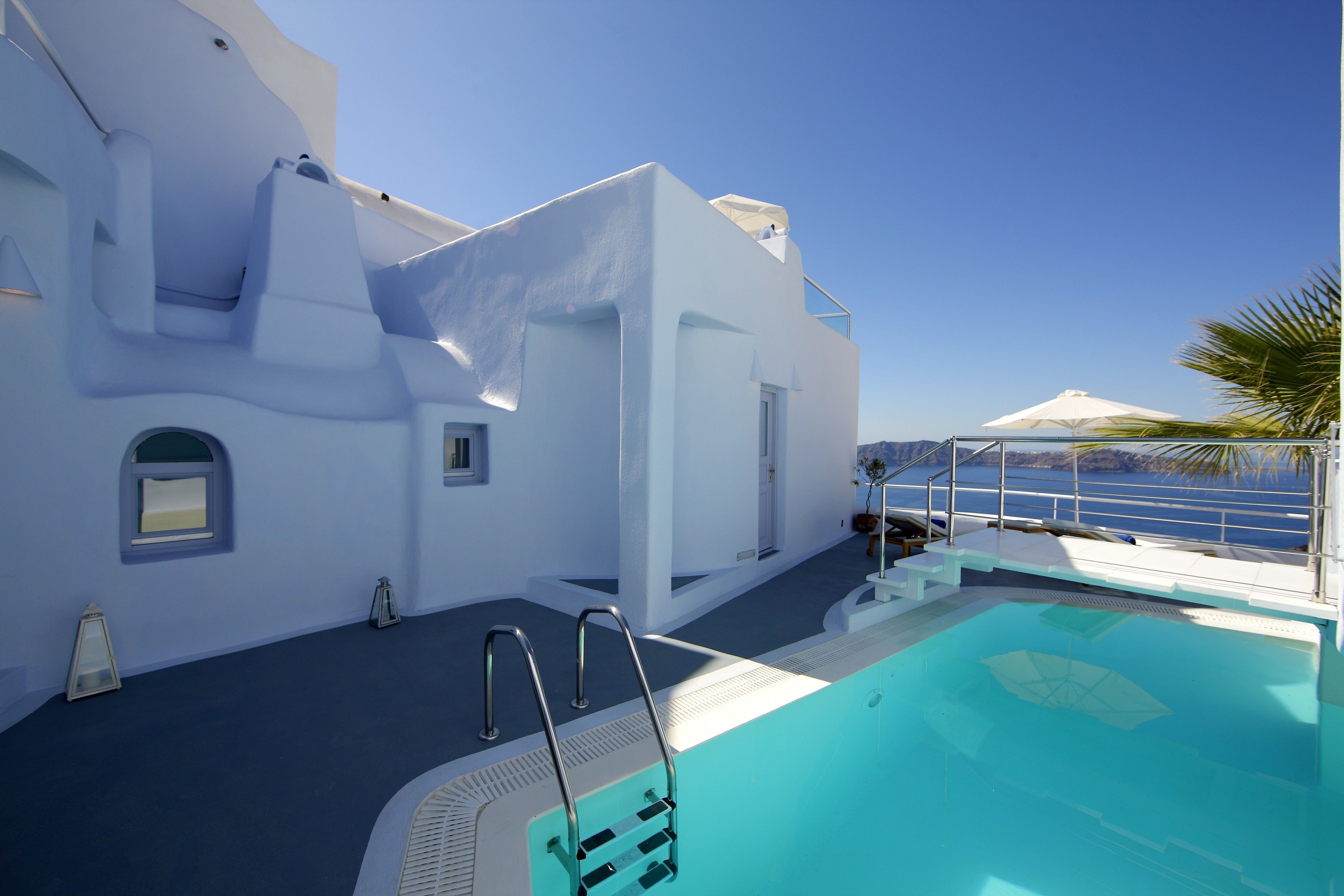 A pool area with a clear view of the sea, a white building with a balcony, and a palm tree.