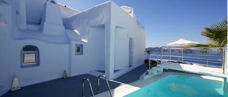 A pool area with a clear view of the sea, a white building with a balcony, and a palm tree.