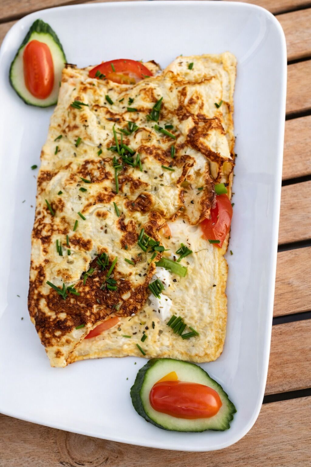 Une assiette rectangulaire blanche sur laquelle est servie une omelette repliée, garnie de ciboulette, accompagnée d’avocat et de tomates en tranches.