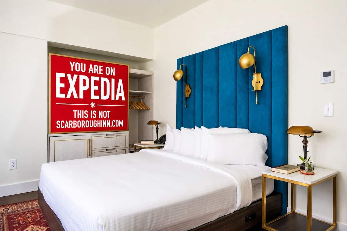 Une chambre d’hôtel avec un grand lit, une tête de lit bleue, des lampes dorées, une table de chevet avec une lampe et un panneau rouge accroché au mur.