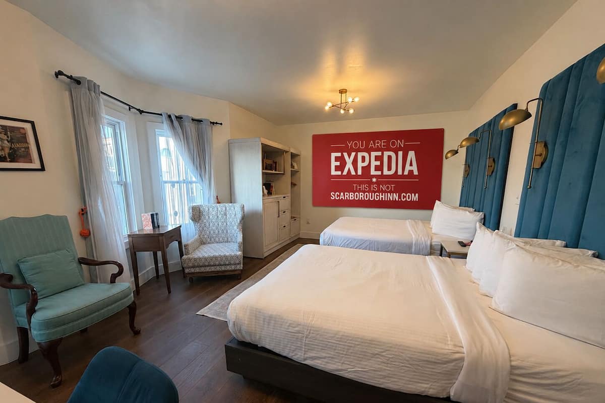 Une chambre d’hôtel avec deux lits, un bureau, une chaise, une armoire et un panneau indiquant que la chambre est disponible sur Expedia.