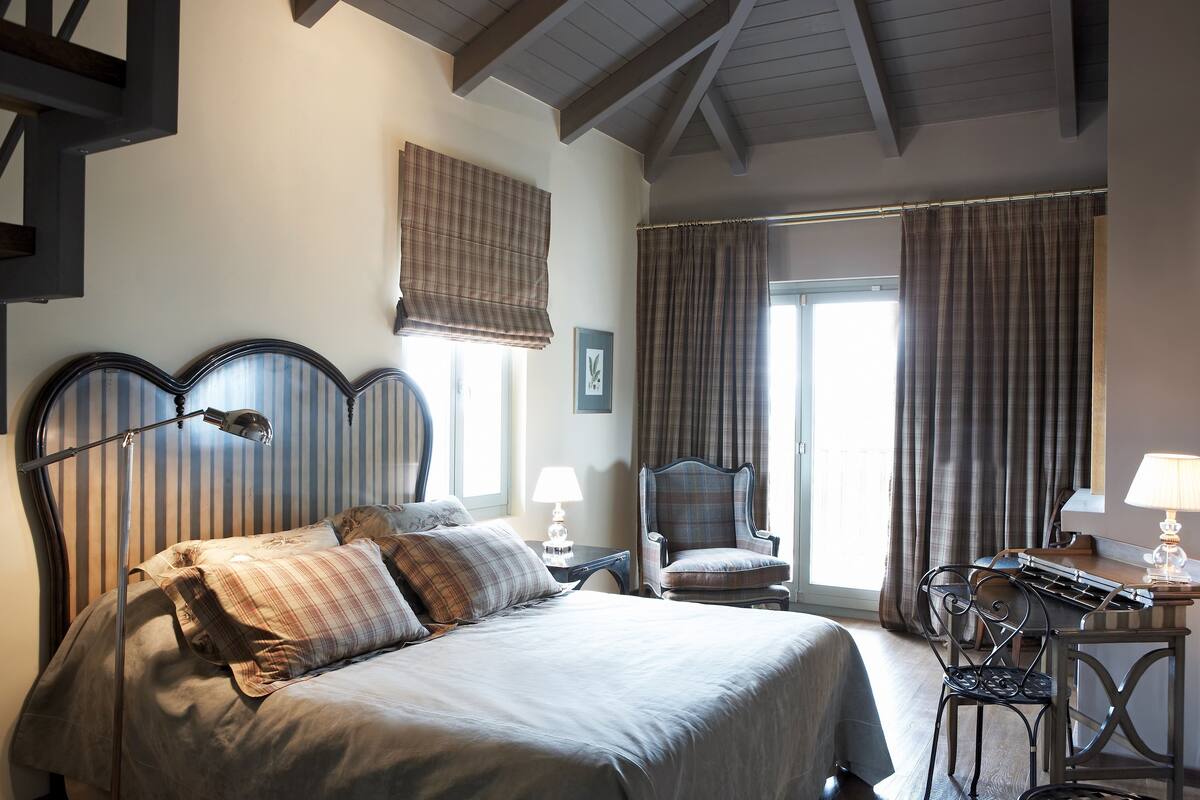 Ein Schlafzimmer mit einem großen Bett, einem Schreibtisch und einem Stuhl. Ein Fenster mit Vorhängen und ein Bild an der Wand sind ebenfalls vorhanden.